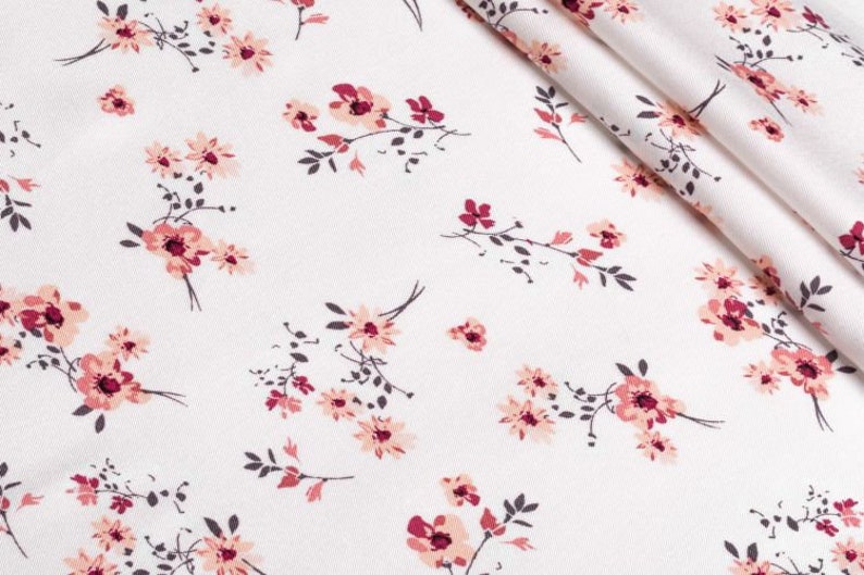 K&ouml;nnte beinhalten: Wei&szlig;er Stoff mit einem sich wiederholenden Blumenmuster. Das Design zeigt Gruppen von rosa und burgunderfarbenen Blumen mit dunkelgr&uuml;nen Stielen und Bl&auml;ttern. Der Stoff ist gefaltet und zeigt die Textur und das Muster.