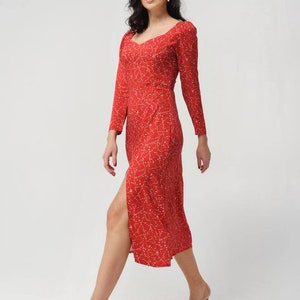 May include: A woman wearing a red midi dress with a white floral pattern and a side slit. She is wearing nude heels and has her hair pulled back.