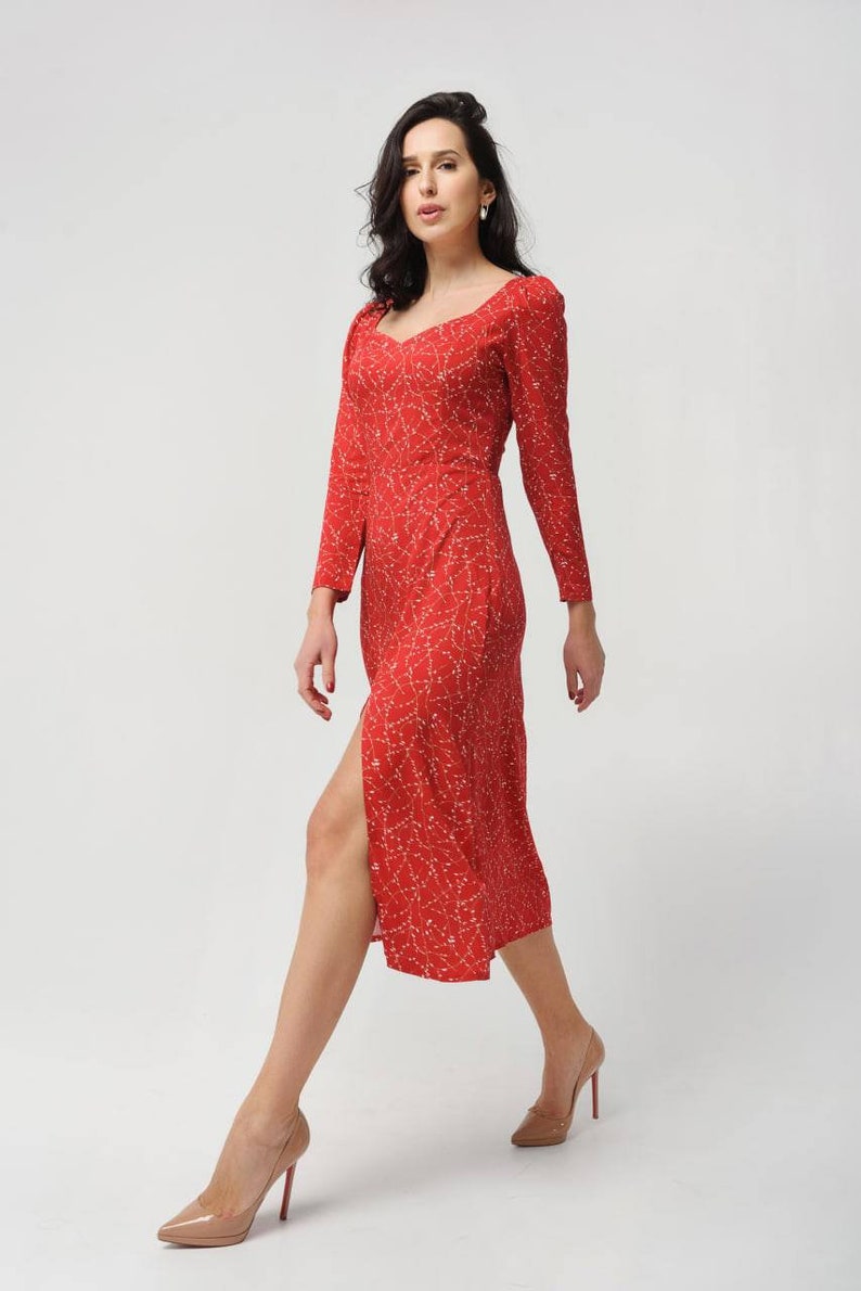 May include: A woman wearing a red midi dress with a white floral pattern and a side slit. She is wearing nude heels and has her hair pulled back.