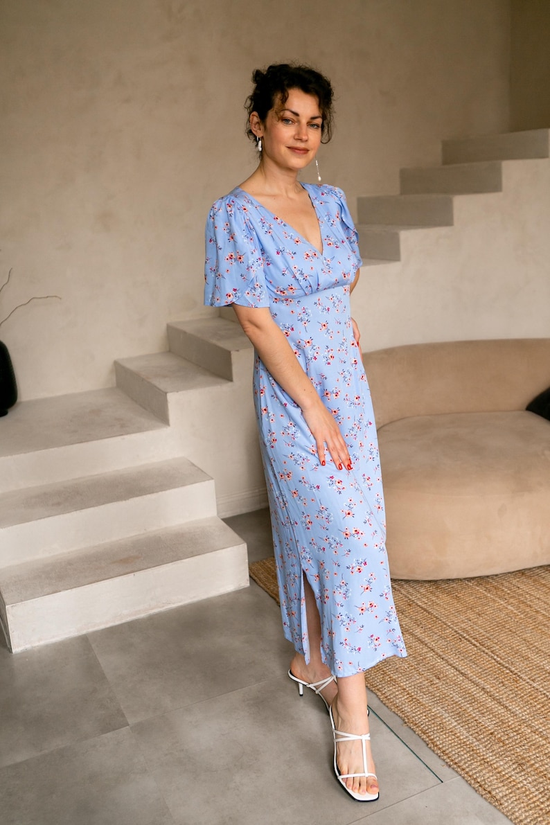 K&ouml;nnte beinhalten: Hellblaues Kleid mit floralem Muster, V-Ausschnitt und kurzen &Auml;rmeln. Das Kleid hat einen Seitenschlitz und wird mit wei&szlig;en Sandalen getragen. Das Blumenmuster besteht aus kleinen roten und wei&szlig;en Bl&uuml;ten.