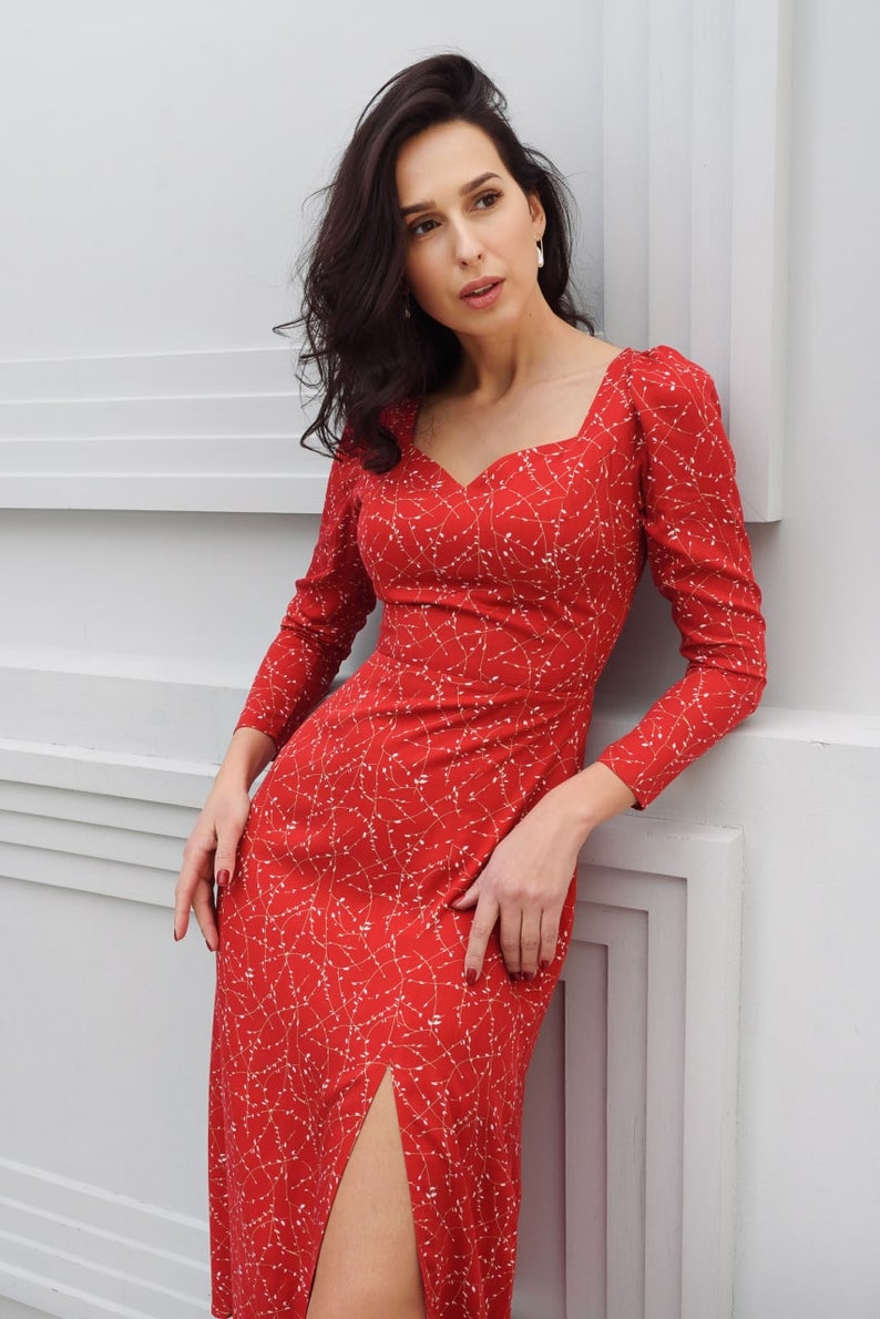 May include: A red midi dress with a white floral pattern and a thigh-high slit. The dress has a sweetheart neckline and long sleeves.
