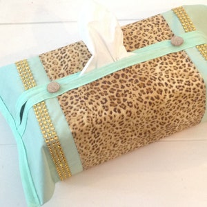 May include: A mint green tissue box cover with a leopard print design and gold trim. The cover has a flap on the top for easy access to tissues.