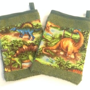 May include: Two green kitchen towels with a dinosaur print. The towels feature a variety of dinosaurs, including a stegosaurus, a brachiosaurus, and a triceratops. The towels are made of a soft, absorbent material and are perfect for drying dishes or hands.