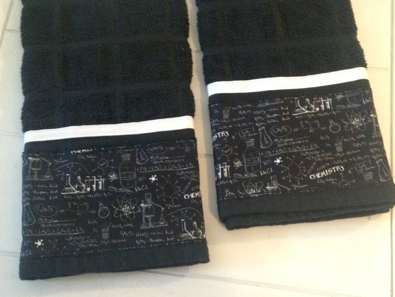May include: Two black hand towels with a white stripe and a decorative band featuring chemistry-themed illustrations and the word "CHEMISTRY". The towels are made of a soft, absorbent material.