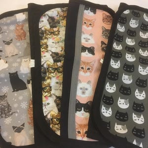 May include: Four black oven mitts with cat-themed designs. One has cats wearing glasses, another a pink background with cat faces, and a third a grey background with cat silhouettes. The fourth mitt has a grey background with cats and snowflakes.