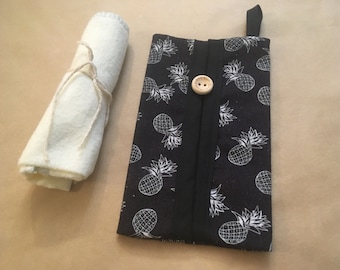May include: A black fabric tissue holder with a white pineapple pattern and a wooden button closure. The holder is rolled up and tied with twine.