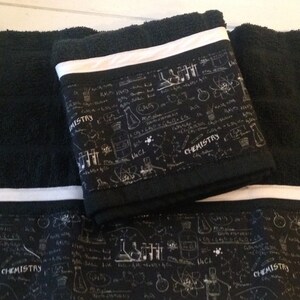 May include: Black bath towels with a white stripe and a decorative band featuring chemistry-themed illustrations and the word "CHEMISTRY" in white. The towels are folded and stacked, showcasing the design.