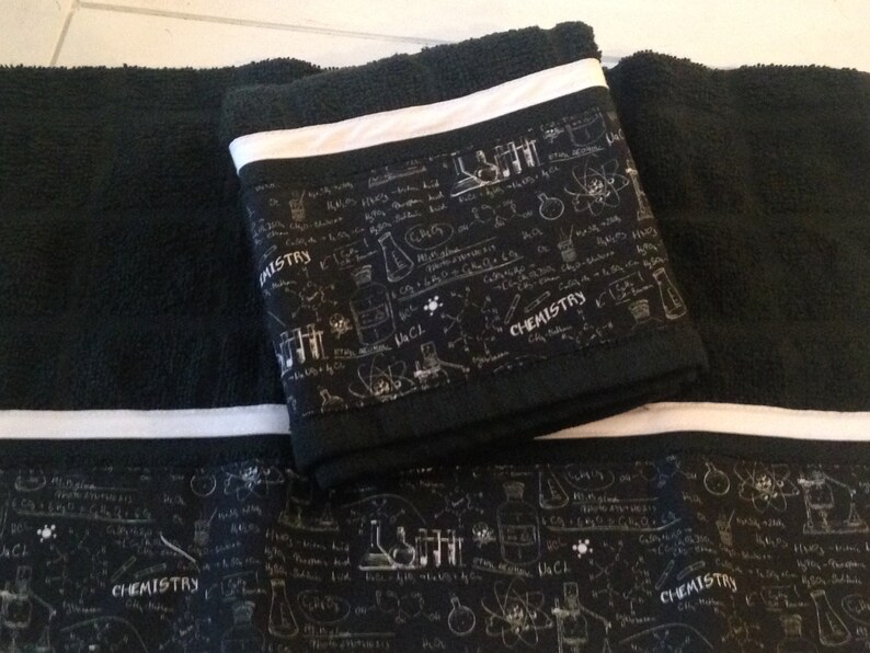 May include: Black bath towels with a white stripe and a decorative band featuring chemistry-themed illustrations and the word "CHEMISTRY" in white. The towels are folded and stacked, showcasing the design.