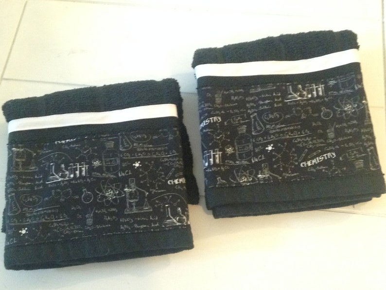 May include: Two black hand towels with a white stripe and a science-themed border. The border features chemistry-related illustrations and the word "CHEMISTRY" in white. The towels are folded.