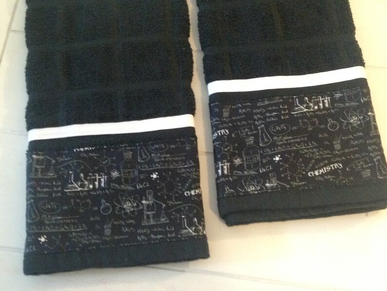 May include: Two black hand towels with a white stripe and a decorative band featuring chemistry-themed illustrations in white. The towels are made of a soft, absorbent material, perfect for the bathroom or kitchen.
