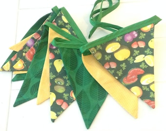 May include: A green and yellow fabric banner with a fruit and leaf pattern. The banner is made of triangular flags with a green leaf pattern on one side and a yellow polka dot pattern on the other side. The flags are sewn together with green ribbon.
