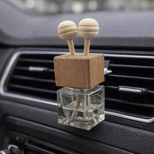 Peut inclure: Un diffuseur de parfum pour voiture avec un flacon en verre transparent et un dessus en cube de bois. Deux sphères en bois de couleur claire sont fixées au sommet. Le diffuseur est placé sur le tableau de bord d'une voiture, près de la grille d'aération.