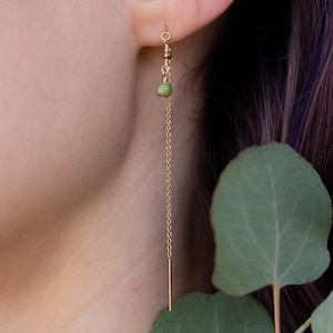 Puede incluir: Un pendiente de color dorado con una cadena y una pequeña piedra verde. El pendiente se lleva en la oreja, con la cadena colgando. El pendiente tiene un diseño sencillo y elegante.