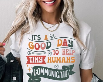 Op de afbeelding: Wit T-shirt met een kleurrijke afbeelding met de tekst "It's a good day to help tiny humans communicate".