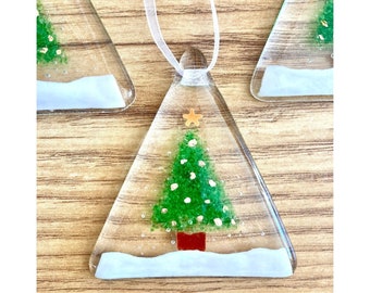 Könnte beinhalten: Ein gläserner Weihnachtsbaumschmuck in Form eines Dreiecks mit einem grünen Weihnachtsbaum im Inneren, einer roten Basis und einem goldenen Stern oben. Der Schmuck befindet sich auf einer Holzoberfläche mit weißem Schnee um die Basis des Baumes.