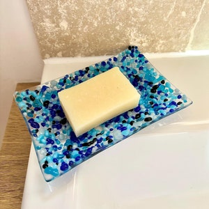 May include: A rectangular soap dish with a mosaic design of blue, light blue, and black glass pebbles. A rectangular bar of light-colored soap rests on the dish. The dish is on a white surface.