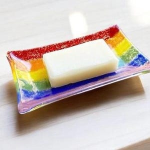 May include: A rectangular glass soap dish with a rainbow design. The dish is made of clear glass with a rainbow pattern on the bottom. A white bar of soap is sitting on the dish.