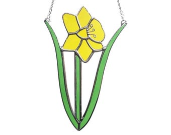 May include: A stained glass daffodil suncatcher with a yellow flower and green leaves. The flower has a detailed center and is framed by a green stem and leaves. A silver chain is attached for hanging.