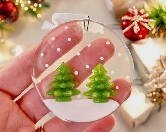 Könnte beinhalten: Ein transparenter Weihnachtsbaumschmuck aus Glas mit weißem Hintergrund und zwei grünen Weihnachtsbäumen. Der Schmuck ist mit weißen Punkten verziert, um Schnee darzustellen.