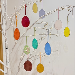 Peut inclure: Une collection de dix œufs de Pâques en verre coloré suspendus à une branche blanche. Les œufs sont dans différentes nuances de jaune, rouge, bleu, vert et rose.