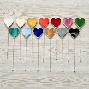 May include: A selection of heart-shaped glass ornaments in a range of colours, such as white, yellow, red, pink, and green. Each heart features a silver-coloured border and a long, twisted silver wire stem. The ornaments are displayed on a light wood-grain surface.