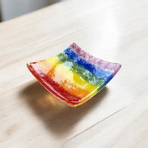 May include: A small, square glass dish with a rainbow design. The dish is made of clear glass with a rainbow of colours embedded in the glass. The colours are red, orange, yellow, green, blue, indigo, and violet.