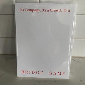 May include: A white card game set in a clear plastic wrapper. The text "2's Company, 3's a Crowd, 4's a" is printed in red at the top, with "BRIDGE GAME" in red at the bottom. The background is a light-colored tile.