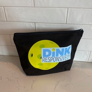 May include: Black canvas zippered pouch with a yellow and blue pickleball graphic and the text "DINK RESPONSIVELY".