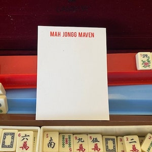 May include: A Mah Jongg game set with white tiles featuring Chinese characters and symbols. The tiles are arranged in rows and columns, with some tiles showing the number "6" and the word "萬". The game set is in a wooden box with a red and blue interior.