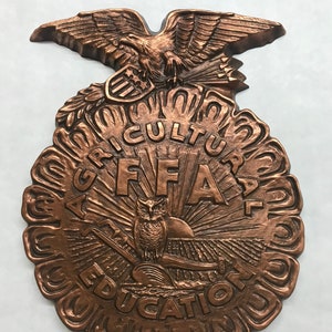 May include: A bronze-colored metal plaque with the Future Farmers of America (FFA) logo. The logo features an eagle with outstretched wings, a shield with stars, an owl, a rising sun, and the words "Agricultural Education" around the outside.