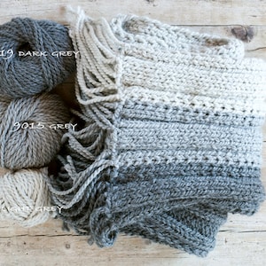 May include: A hand-knitted scarf in shades of gray, with fringe detail, and three balls of yarn in coordinating colors. The yarn colors are labeled: 0519 Dark Grey, 9015 Grey, and 9020 Light Grey. The scarf is folded on a wooden surface.