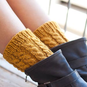 Puede incluir: Un par de polainas de punto de color amarillo mostaza con un patrón de punto de cable. Las polainas se llevan sobre botas de cuero negro.