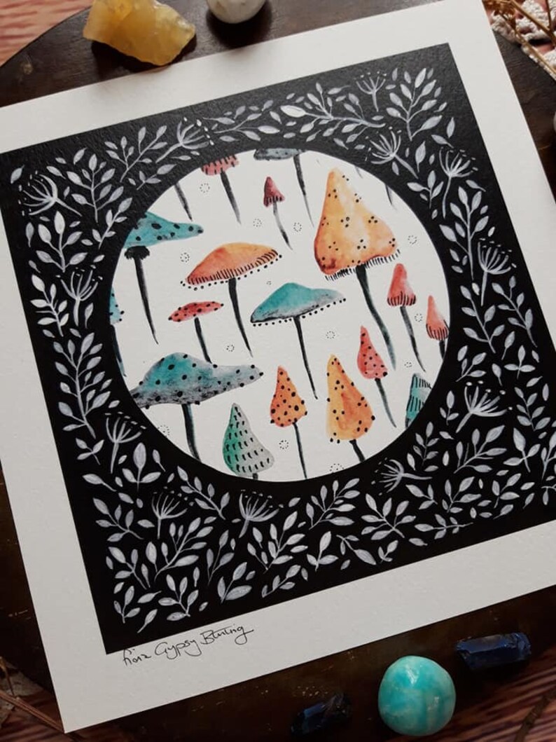 May include: A black and white illustration of a circle filled with colorful mushrooms. The circle is surrounded by a black frame with white leaves and branches. The artist's signature, "Ann Gypsy Bunting", is in the bottom left corner.