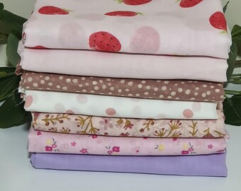 May include: A stack of fabric pieces in various patterns and colors. The top layer features a light pink fabric with red strawberries. Other fabrics include polka dots, floral prints, and solid colors like lavender. Ideal for sewing and crafting projects.