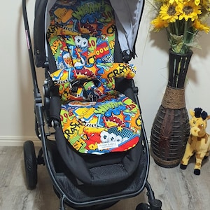 May include: A black baby stroller with a colorful comic book-style patterned seat and canopy. The seat features words like "WHAM!" and "POW!" in a vibrant mix of colors. The stroller has a black frame and wheels.