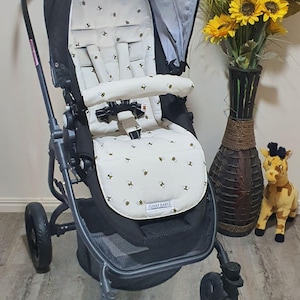 May include: A black and gray stroller with a white seat cover patterned with yellow bees. The stroller has a gray canopy and black wheels. A vase of sunflowers and a stuffed giraffe are in the background. The text "FUNKY BABYZ" is on the seat.