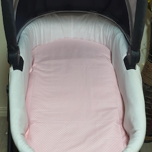 May include: A baby stroller bassinet with a pink polka dot patterned mattress. The bassinet has a white interior and a black exterior frame and canopy. The stroller is designed for infant use.