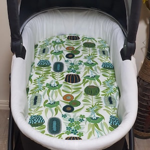May include: A baby stroller bassinet with a white interior and a patterned green, blue, and orange floral print fitted sheet. The stroller has a black frame and a black canopy.