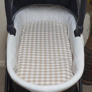 May include: A baby stroller interior with a beige and white gingham patterned mattress pad. The stroller's interior is white, and the exterior is black. The stroller is empty and ready for use.