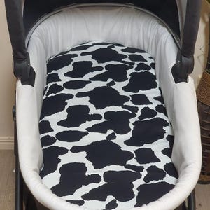 May include: A baby stroller bassinet with a black and white cow print mattress pad. The bassinet is white with a black frame and canopy. The cow print pattern is a repeating design of black irregular shapes on a white background.