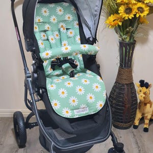 May include: Black stroller with a mint green seat cover patterned with white daisies. The stroller has a black frame, a sunshade, and a safety harness. The words "FUNKY BABYZ" are printed on the seat cover.