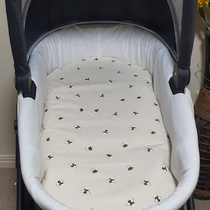 May include: A baby stroller with a cream-colored interior and a patterned bee-themed insert. The insert features a repeating pattern of black and yellow bees on a cream background. The stroller has a black frame and a white interior lining.