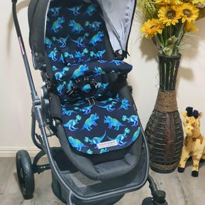 May include: A black stroller with a blue dinosaur pattern on the seat. The stroller has a canopy and a storage basket. The seat is padded and has a five-point harness. The stroller has three wheels and a handle.