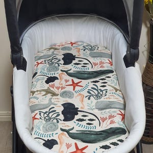 May include: A baby stroller with a white interior and a patterned insert. The insert features a cream background with illustrations of marine life, including whales, seahorses, and starfish in shades of blue, green, and coral.