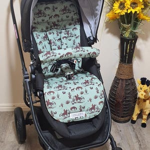 May include: A black stroller with a patterned seat cover featuring cowboys, horses, and cacti on a light green background. The stroller has a black frame, wheels, and a sunshade. The text "FUNKY BABYZ" is visible on the seat.