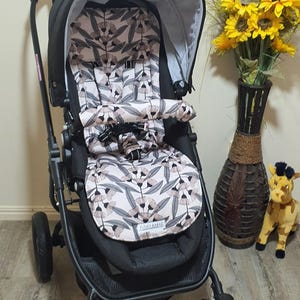 May include: Black stroller with a patterned seat featuring pink and gray floral designs. The stroller has a black frame, wheels, and a canopy. A vase of sunflowers and a stuffed giraffe are in the background.