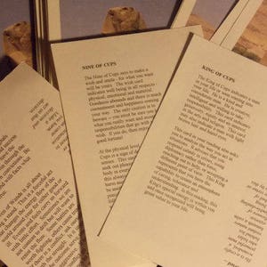 May include: Three off-white rectangular cards with black printed text describing tarot card meanings. The cards are titled Nine of Cups, King of Cups, and Seven of Wands. Each card features a small color image of a desert landscape at the top.