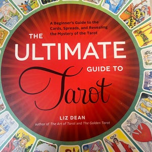 May include: A book titled The Ultimate Guide to Tarot by Liz Dean, featuring a red circular center with white and black text. The cover is surrounded by a border of various colorful tarot card illustrations, including figures, symbols, and a heart.