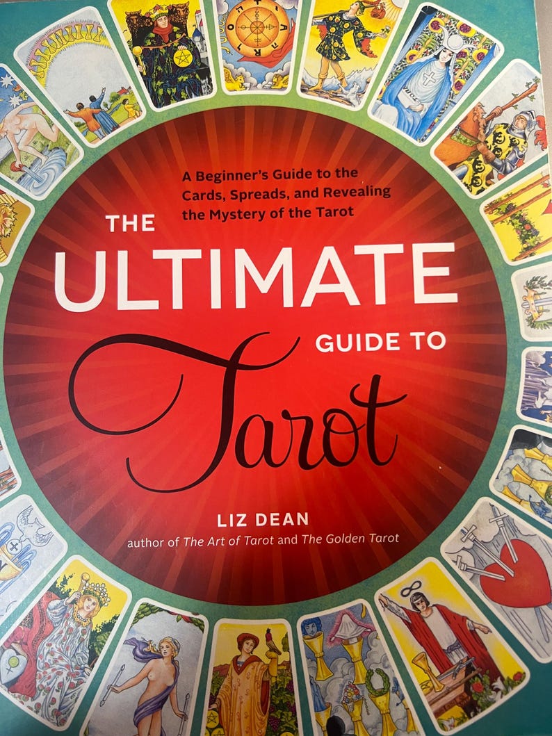 May include: A book titled The Ultimate Guide to Tarot by Liz Dean, featuring a red circular center with white and black text. The cover is surrounded by a border of various colorful tarot card illustrations, including figures, symbols, and a heart.