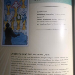 May include: A page from a book featuring the Seven of Cups tarot card illustration. The card shows a dark figure facing seven floating golden cups containing various symbols, including a face, a cloth, jewels, a wreath, and a dragon.
